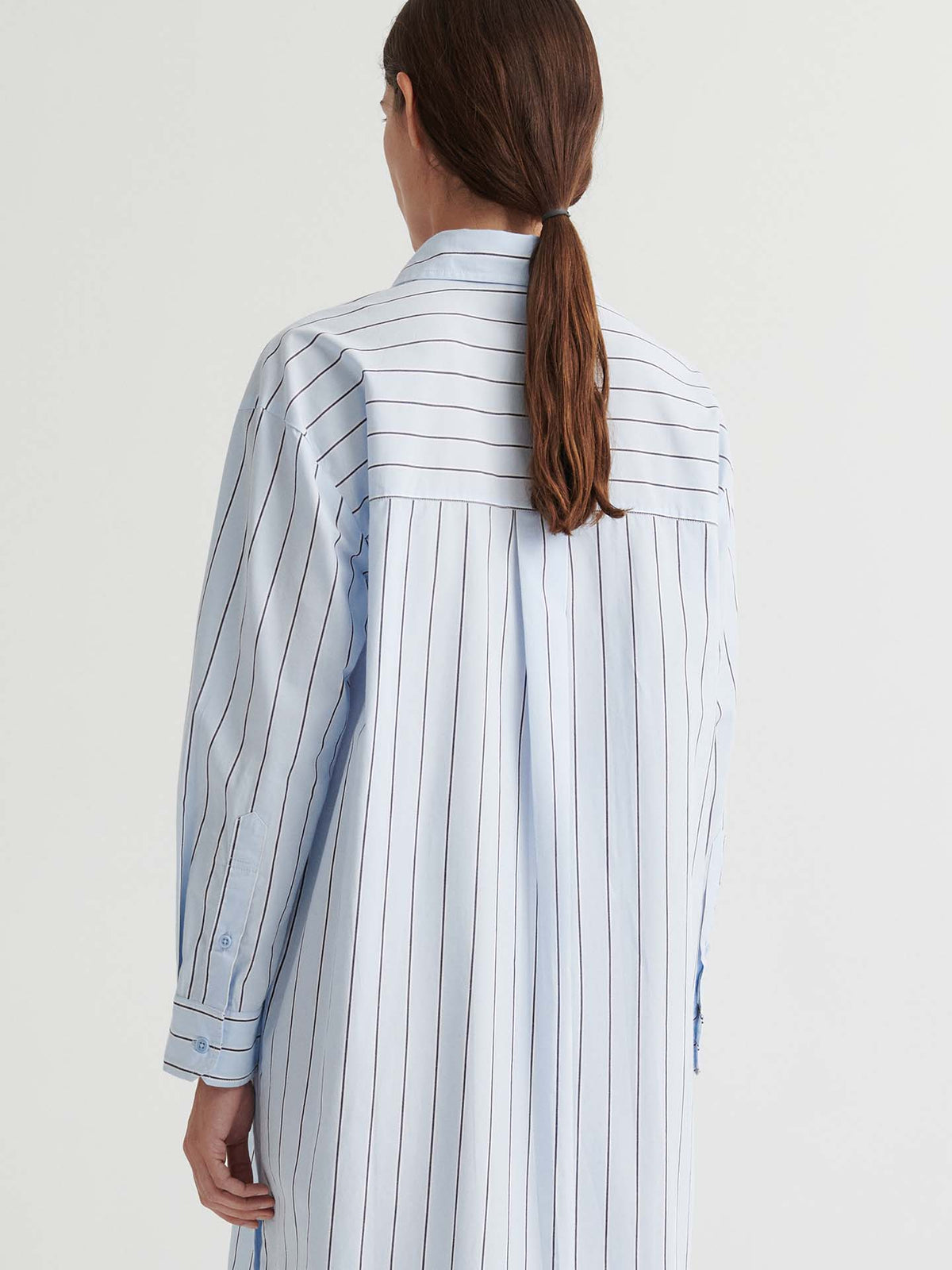 A person with long brown hair is shown from the back against a plain background, wearing the Elementary Dress – Cloud Stripe, featuring light blue and white vertical stripes in a relaxed, boxy fit.