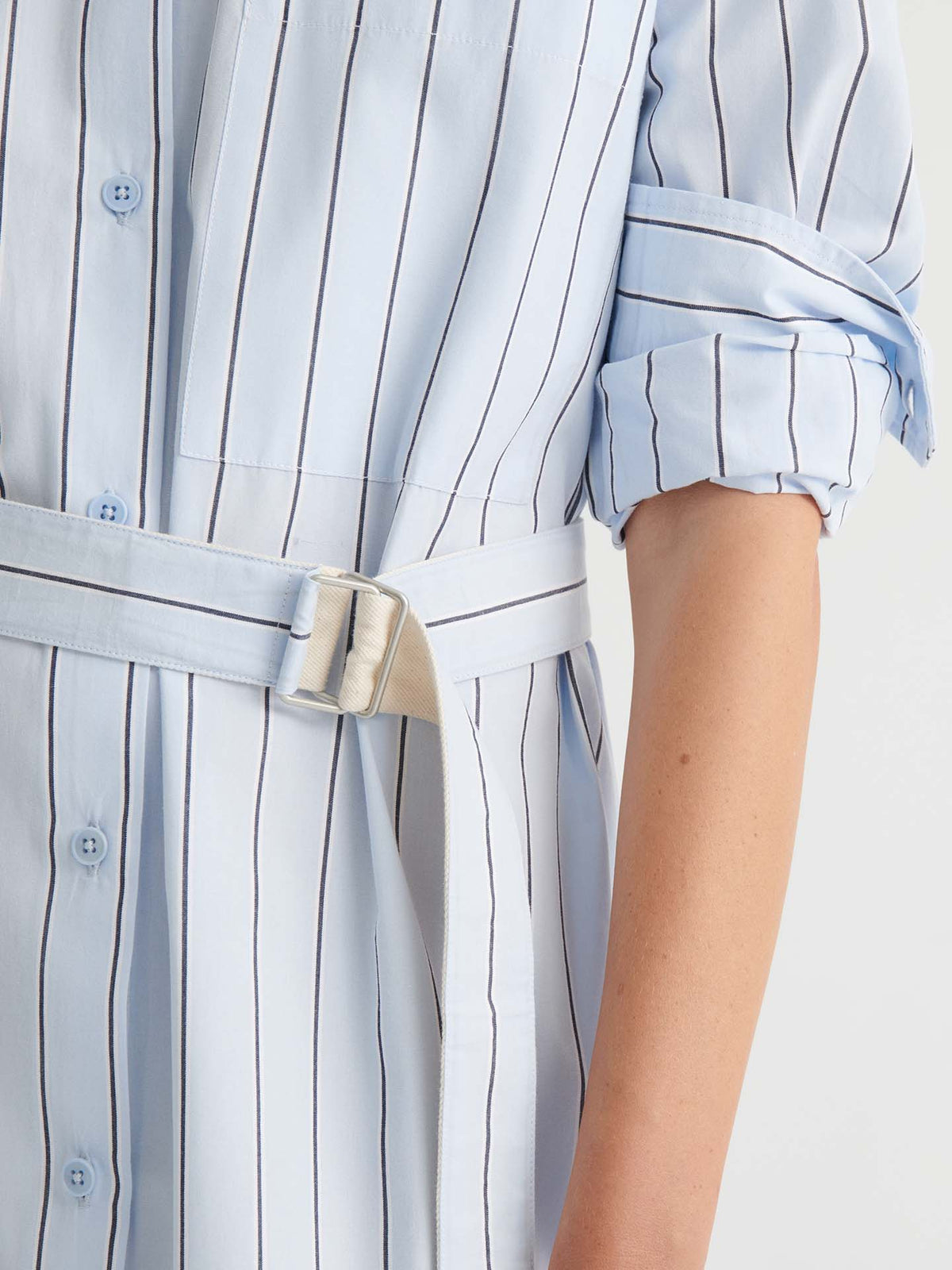 A person wears the Elementary Dress in Cloud Stripe—a light blue, vertically striped shirt dress featuring rolled-up sleeves, a belted waist, and a boxy fit for a relaxed look. Only their torso and one arm are visible.
