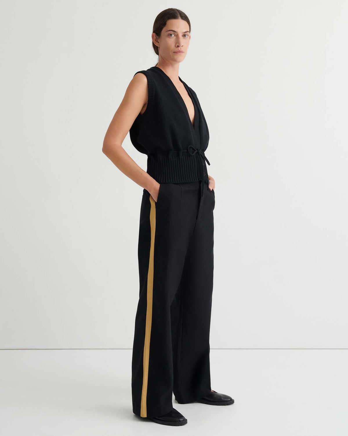 A person stands in a minimal studio wearing the Elworthy Vest – Black styled as a sleeveless top, paired with black wide-leg pants featuring a yellow side stripe and black shoes. Hands in pockets, facing slightly to the left.