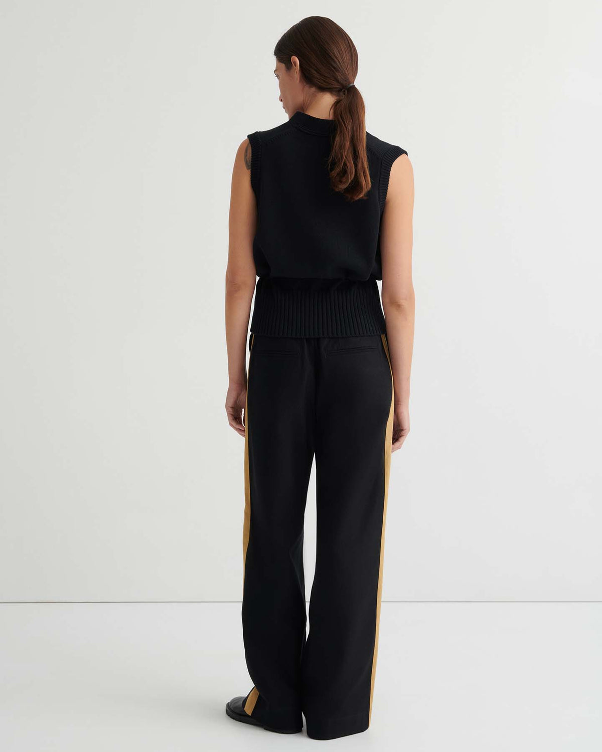 A person stands facing away from the camera, wearing the Elworthy Vest – Black paired with black wide-leg pants featuring yellow side stripes, in a minimalist white setting.