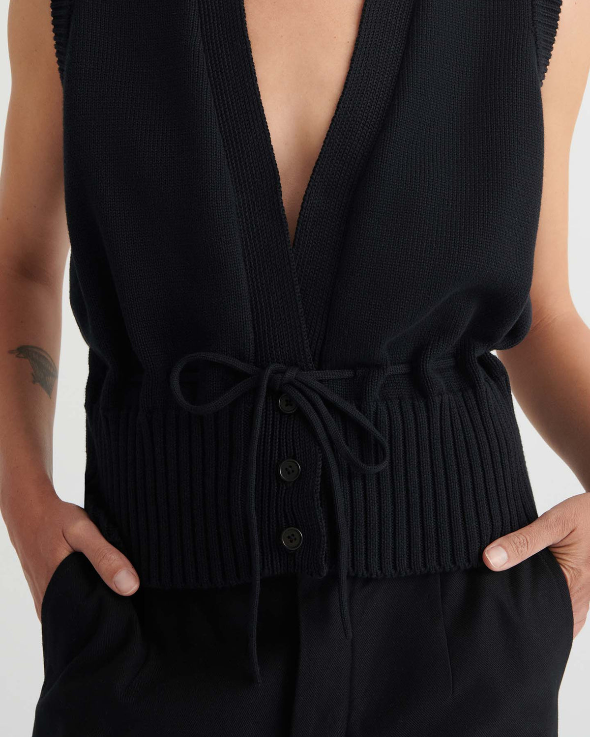 A person wears the Elworthy Vest – Black, a relaxed-fit, sleeveless knit top with a deep V-neck, tie waist, and button details. Their hands are tucked into pockets, revealing a small tattoo on the forearm.