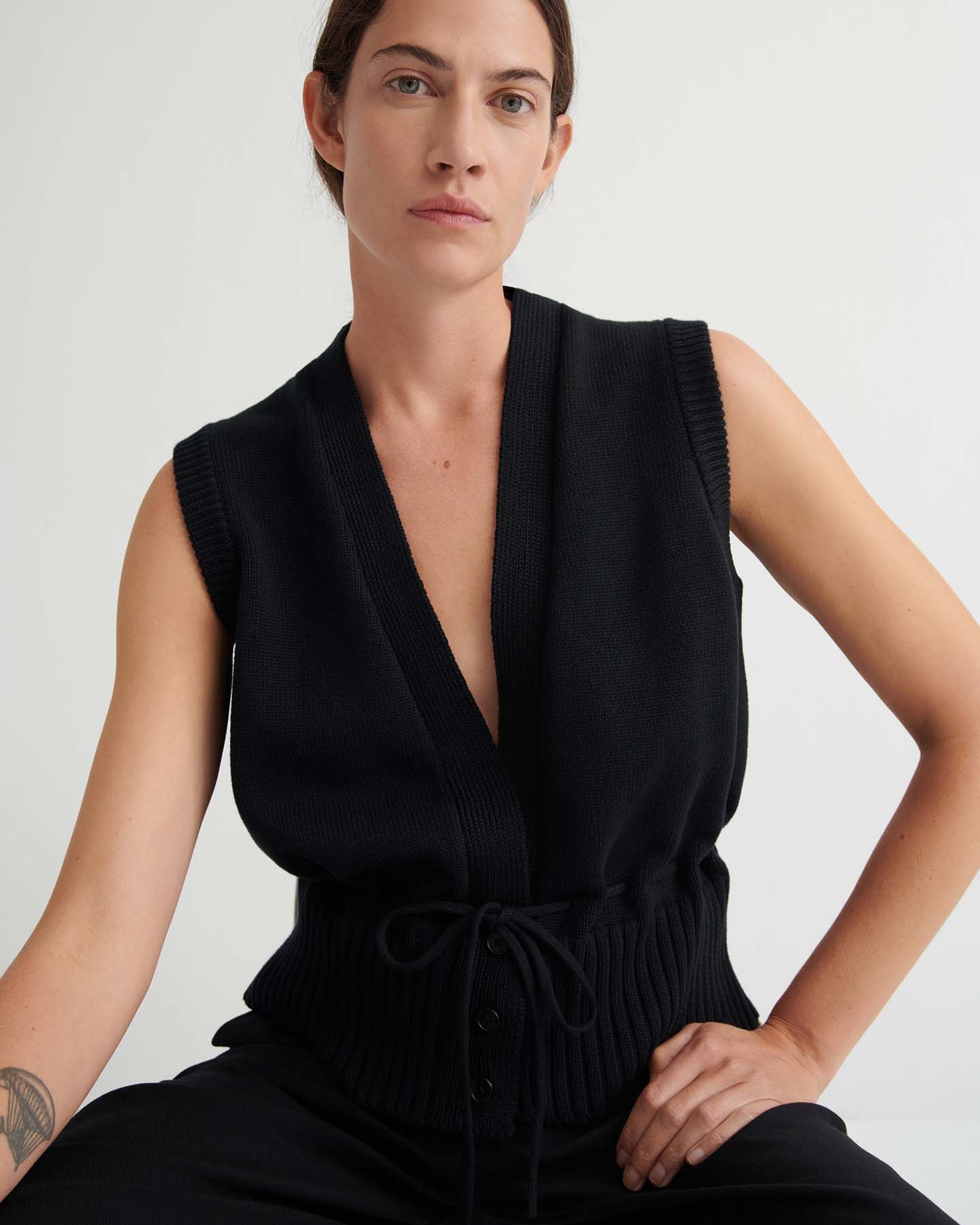 Wearing the Elworthy Vest – Black, a sleeveless knit with a deep V-neck and drawstring waist, the person showcases relaxed fit style while seated against a simple light background.