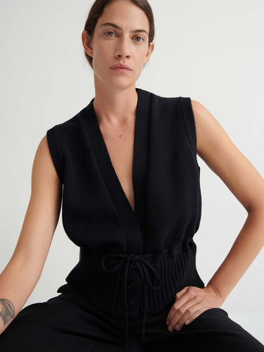 Wearing the Elworthy Vest – Black, a sleeveless knit with a deep V-neck and drawstring waist, the person showcases relaxed fit style while seated against a simple light background.