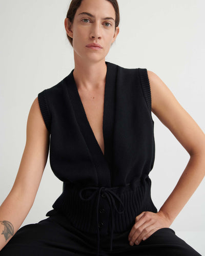 Wearing the Elworthy Vest – Black, a sleeveless knit with a deep V-neck and drawstring waist, the person showcases relaxed fit style while seated against a simple light background.
