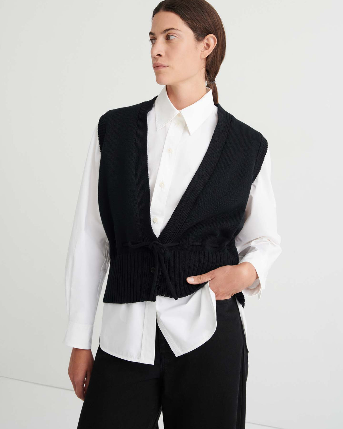 A person stands against a plain light-colored wall, dressed in black pants and a white button-up shirt layered under the relaxed fit Elworthy Vest – Black.