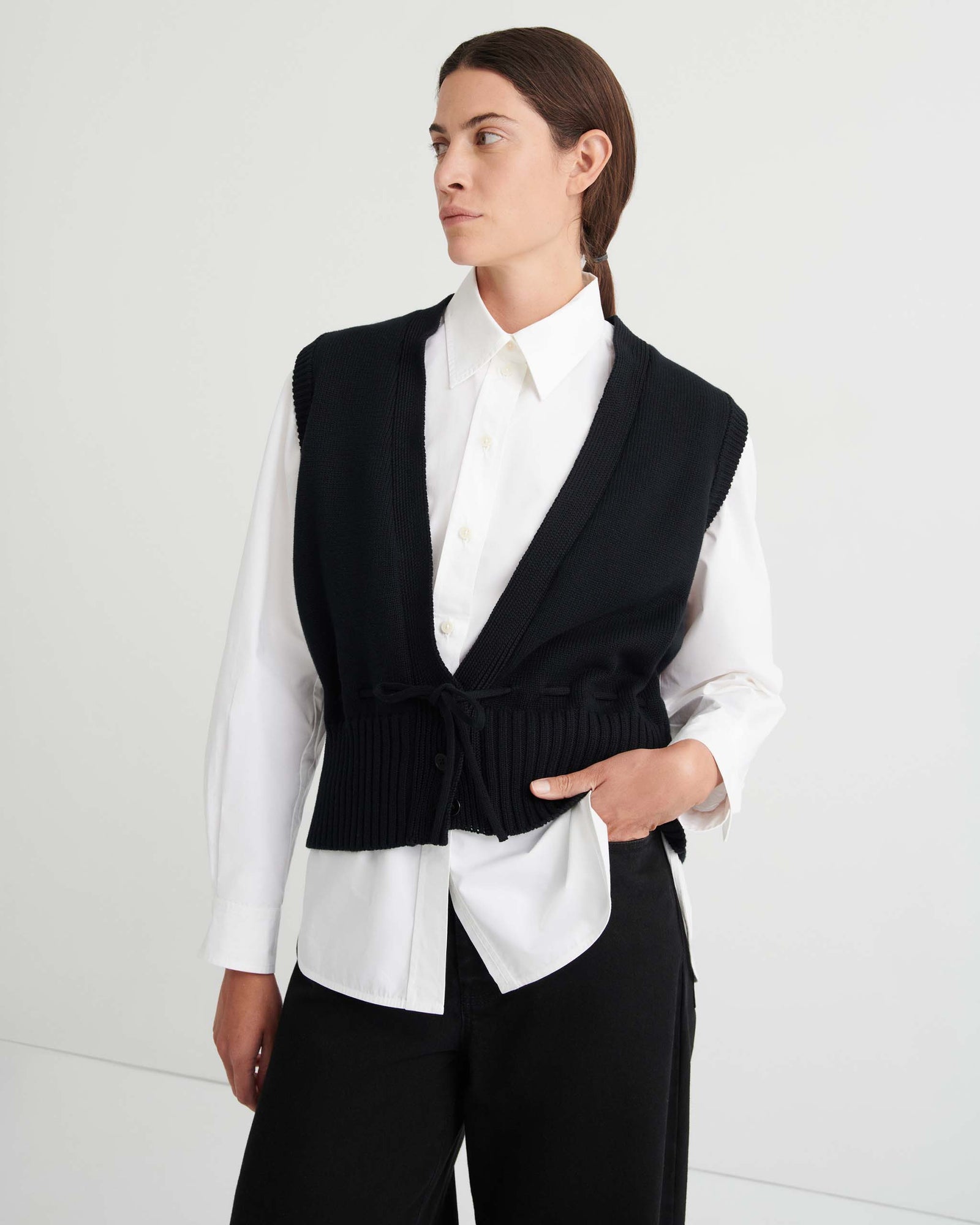 Wearing the Elworthy Vest – Black, a sleeveless knit with a deep V-neck and drawstring waist, the person showcases relaxed fit style while seated against a simple light background.