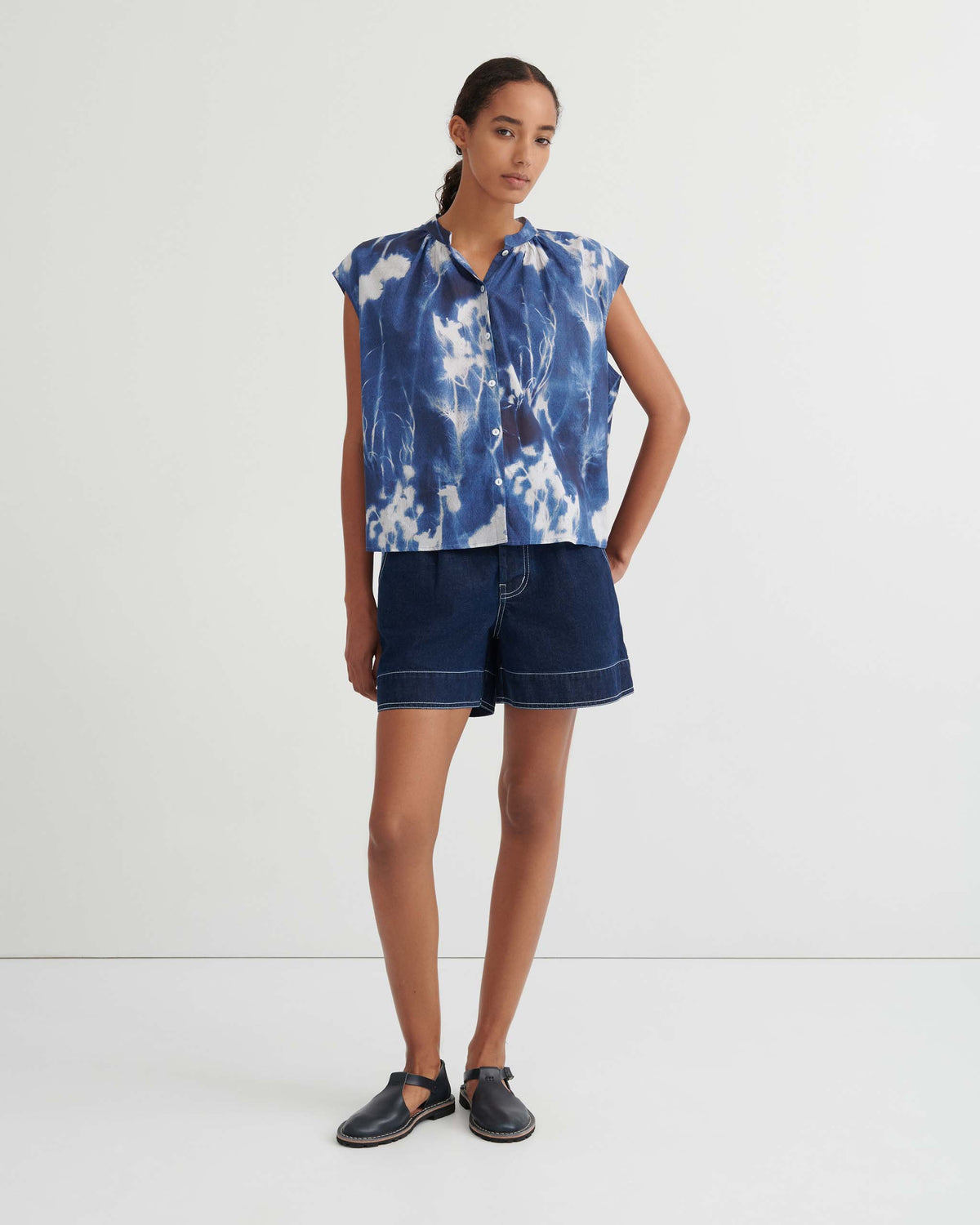 A woman stands against a white wall wearing a blue tie-dye top, dark denim shorts, and black sandals.