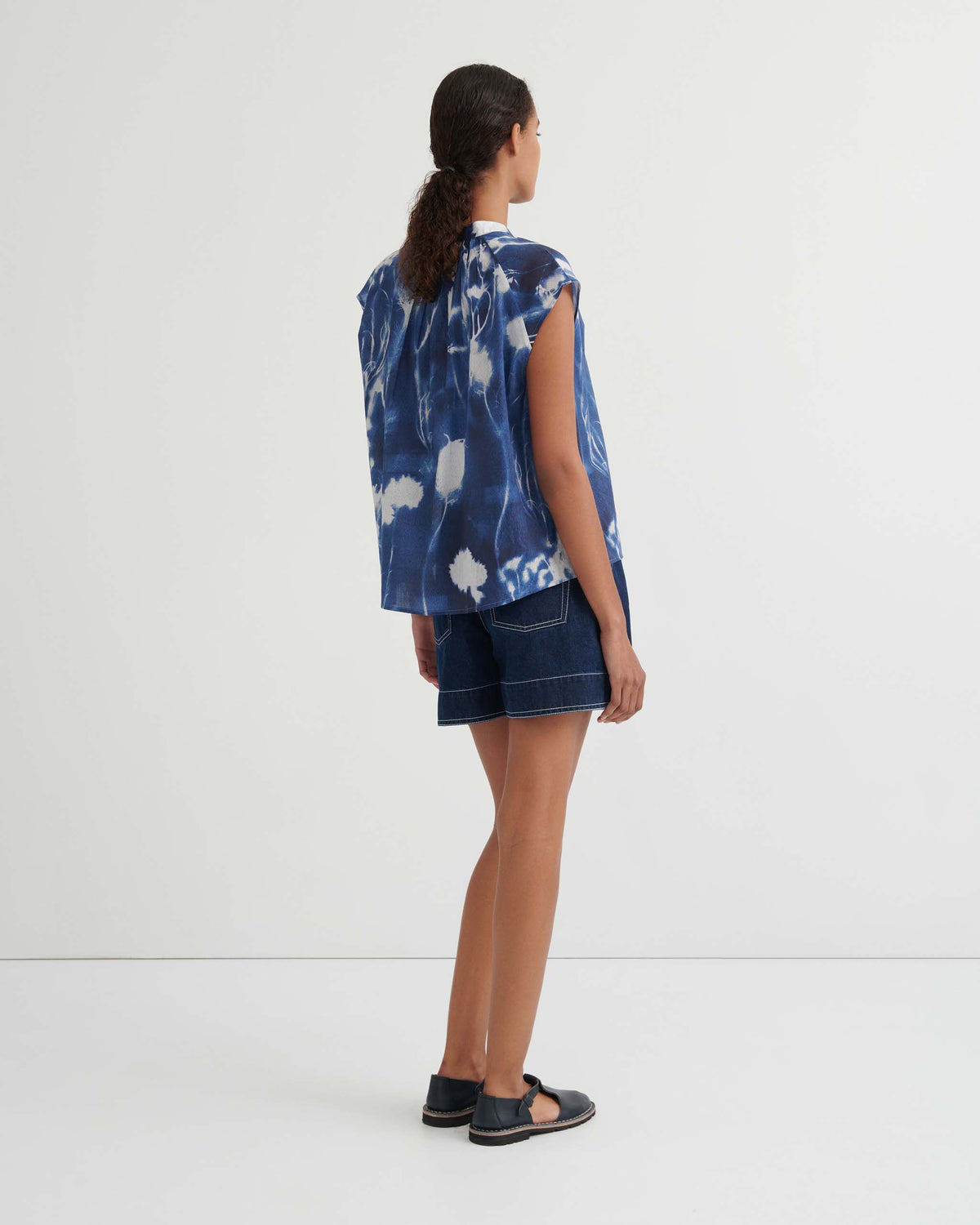 Person standing against a plain white wall, wearing a blue tie-dye sleeveless top, dark denim shorts, and black sandals. Their hair is tied back in a braid.