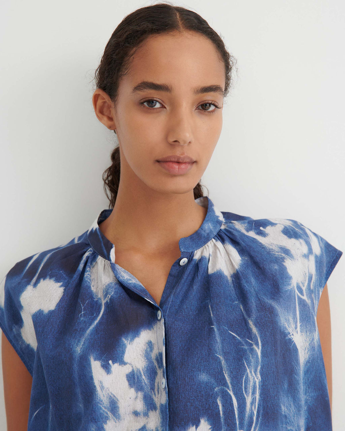 A person with long dark hair in a ponytail wears a blue and white patterned button-up top, standing against a plain light background.