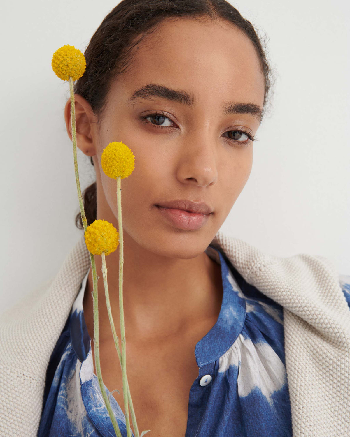 Woman in a blue patterned shirt holds yellow billy ball flowers, looking at the camera against a plain white background.