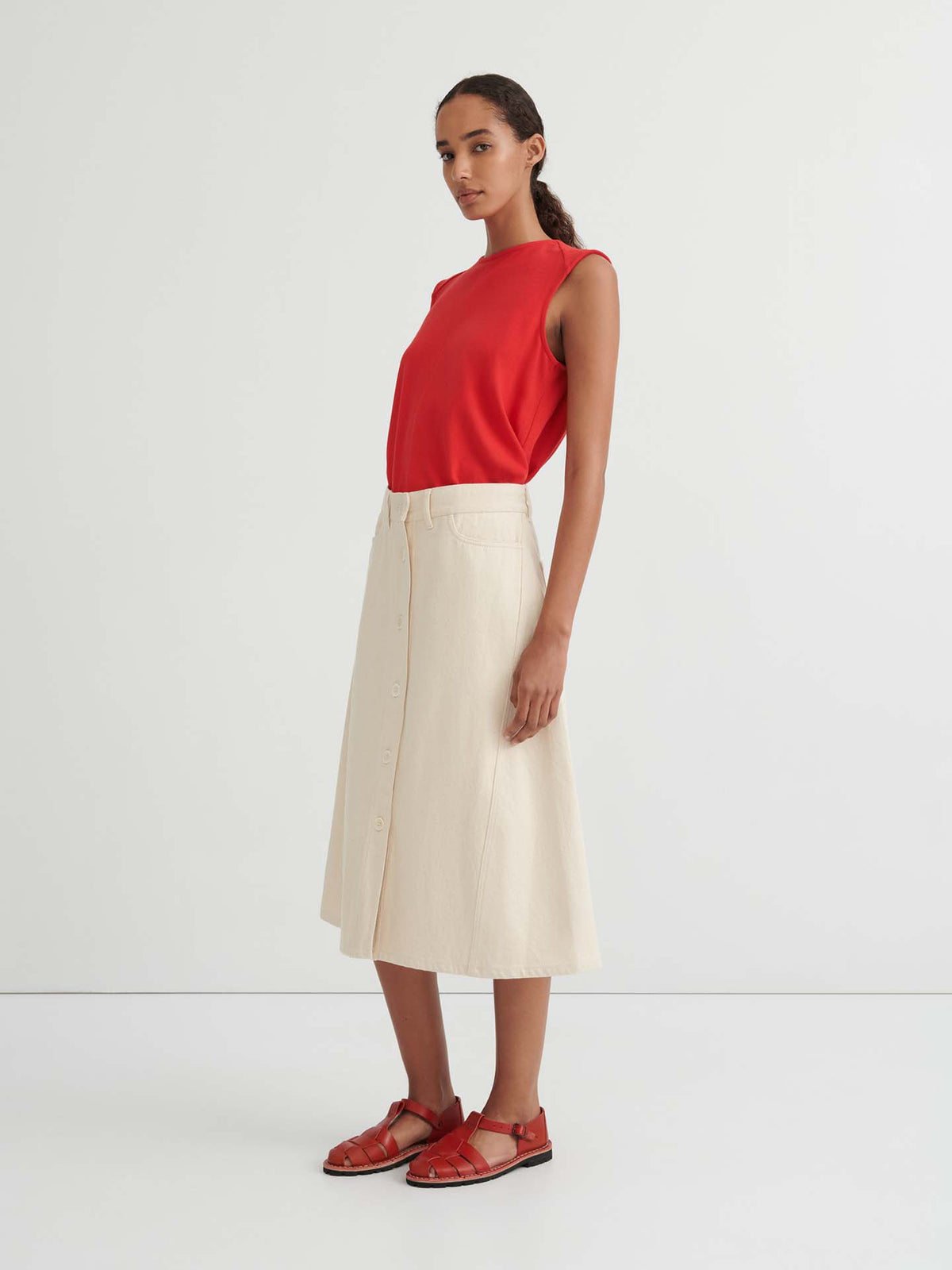 A woman stands against a plain background, wearing a sleeveless red top, the Jubilee Skirt in Ecru denim featuring a button-down design and relaxed fit, and red sandals.