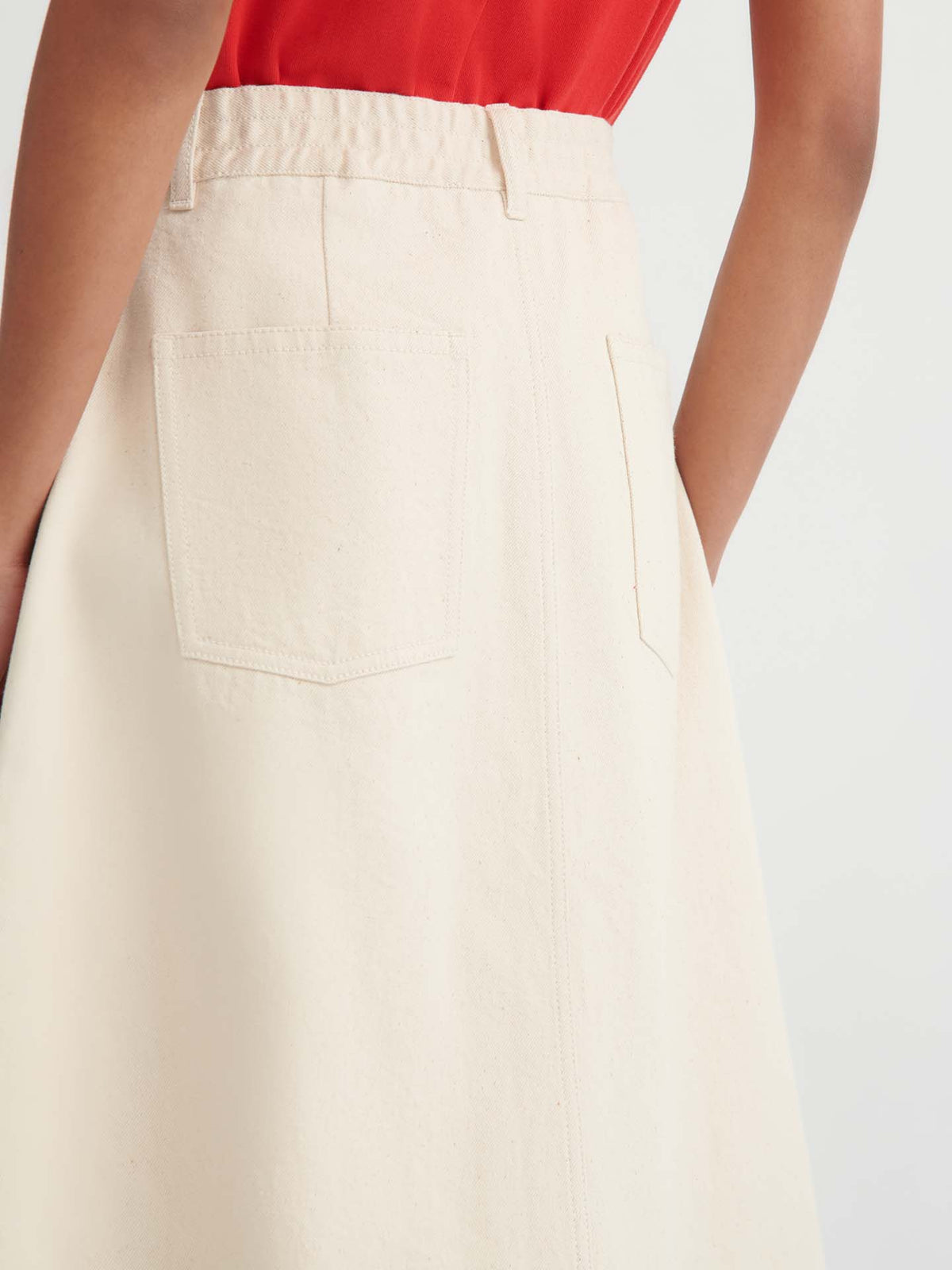 Wearing a sleeveless red top and the Jubilee Skirt – Ecru denim, featuring a high-waisted design with two back pockets, this person stands with hands in pockets, highlighting the skirt’s relaxed fit for an effortlessly chic look.