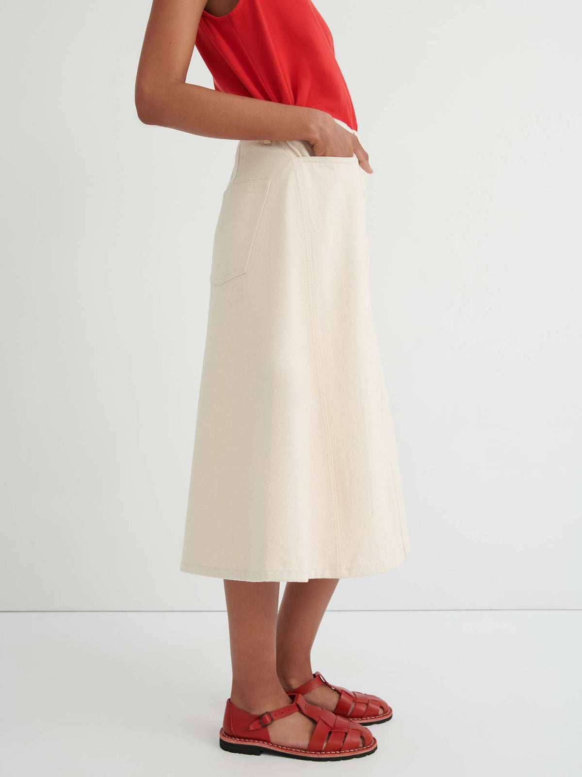 A person wearing a sleeveless red top and the Jubilee Skirt – Ecru denim, a cream midi women’s skirt with pockets and a relaxed fit, paired with red closed-toe sandals, stands against a plain white background.
