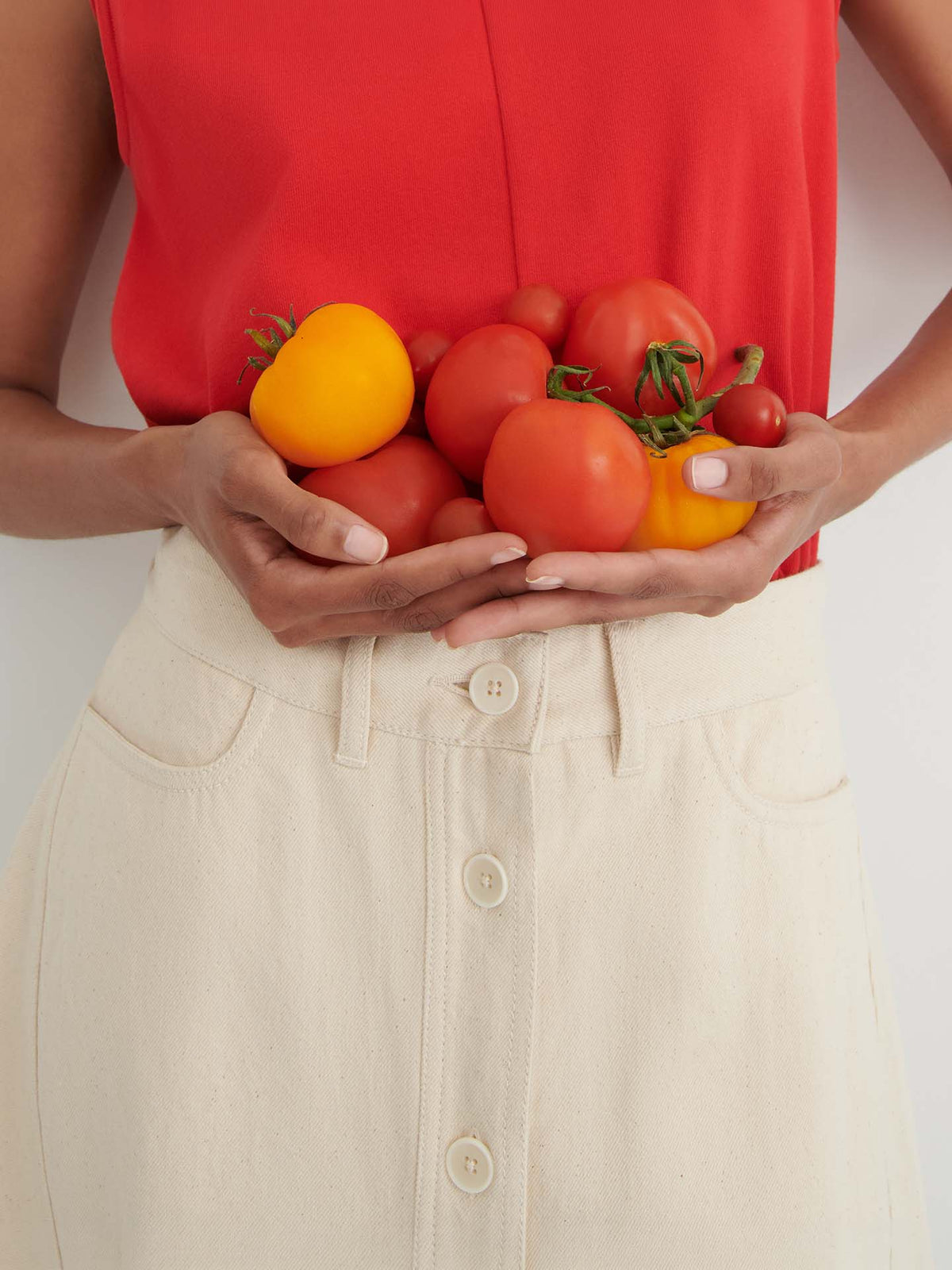 A person in a red top and the Jubilee Skirt – Ecru denim holds several red and yellow tomatoes in their hands against a plain background.