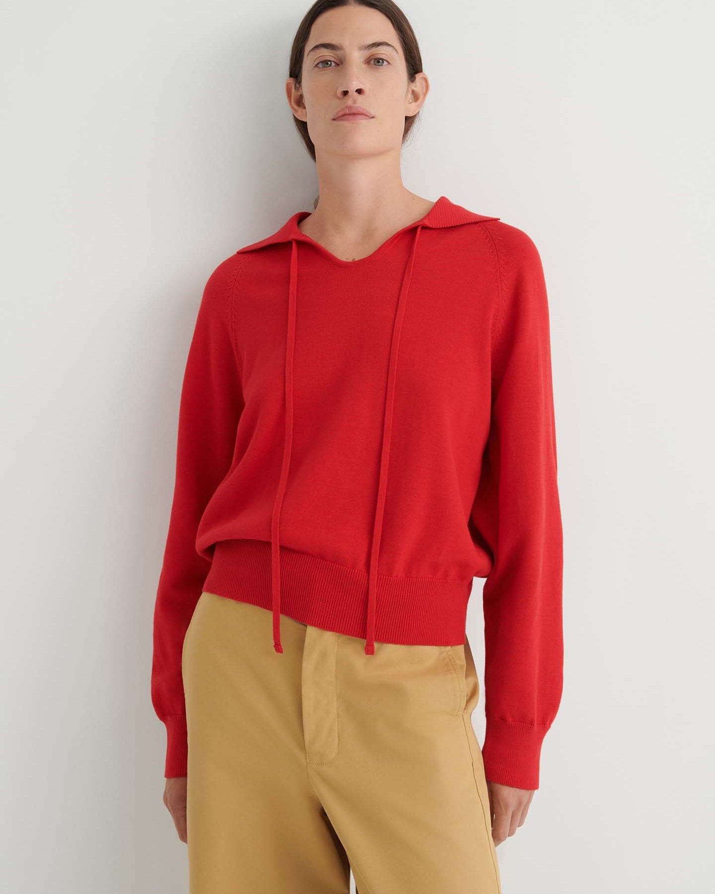 A person wearing the Keyhole Jumper in Poppy Red—a relaxed fit, long-sleeve hoodie with drawstrings—paired with tan pants, stands against a plain white background.