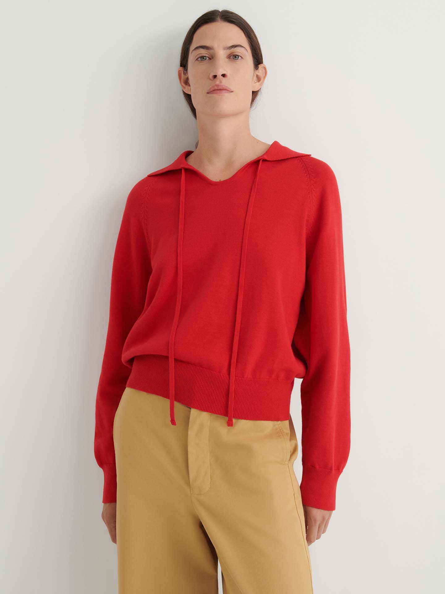 A person wearing the Keyhole Jumper in Poppy Red—a relaxed fit, long-sleeve hoodie with drawstrings—paired with tan pants, stands against a plain white background.