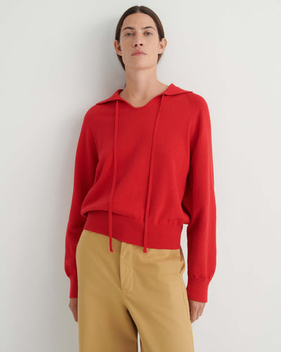 A person wearing the Keyhole Jumper in Poppy Red—a relaxed fit, long-sleeve hoodie with drawstrings—paired with tan pants, stands against a plain white background.