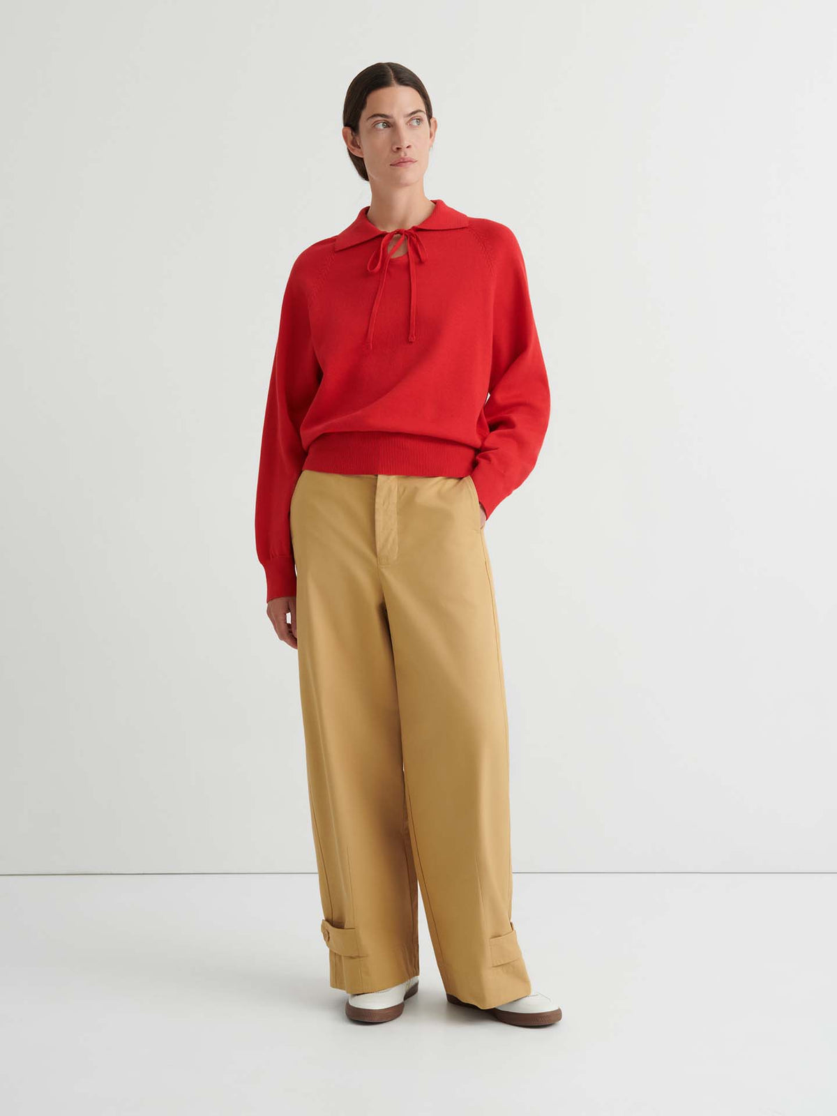 A person stands against a plain white background wearing the Keyhole Jumper in Poppy Red, featuring a relaxed fit with a collar and tie, paired with tan wide-leg cuffed pants and light-colored shoes.