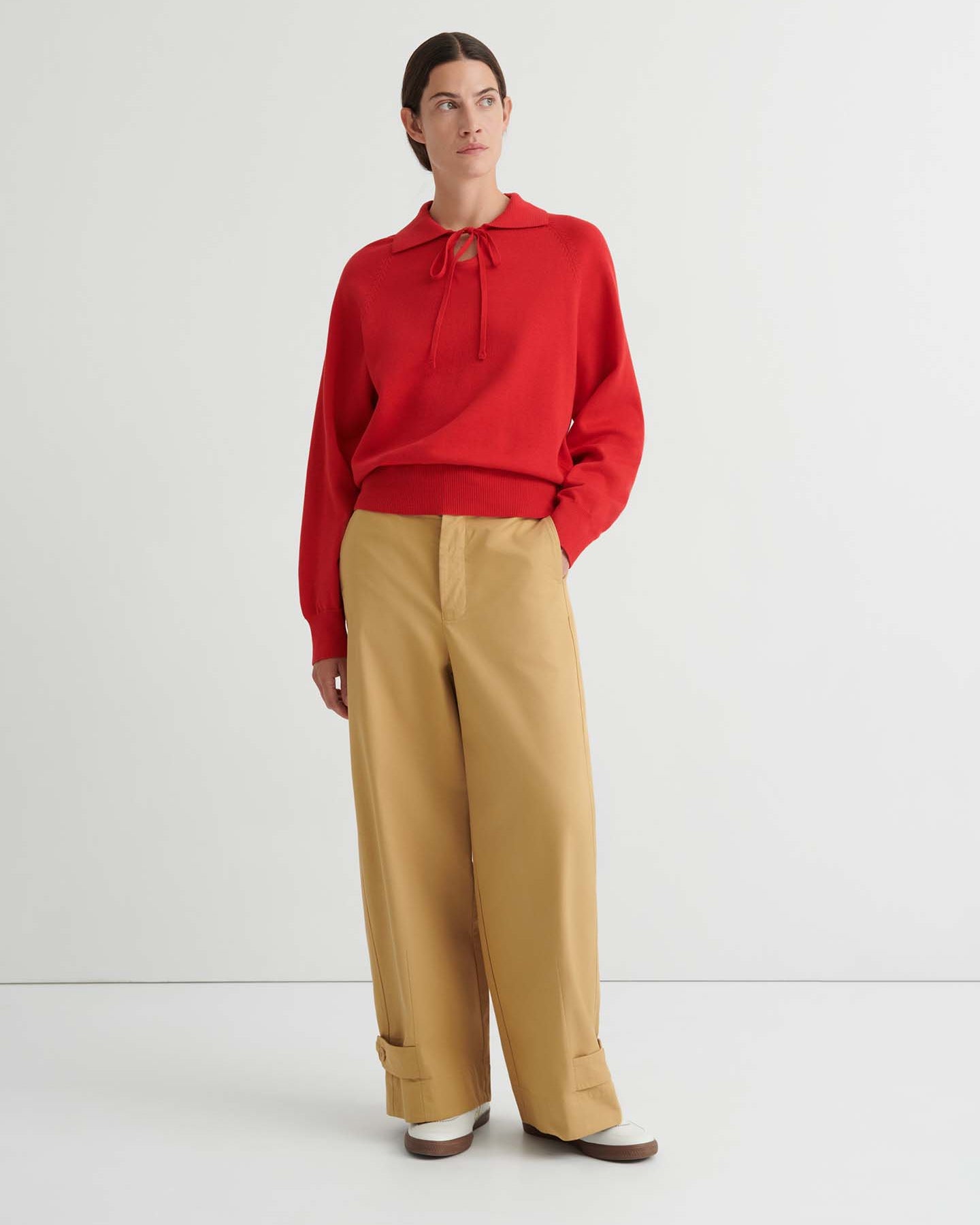 A person wearing the Keyhole Jumper in Poppy Red—a relaxed fit, long-sleeve hoodie with drawstrings—paired with tan pants, stands against a plain white background.