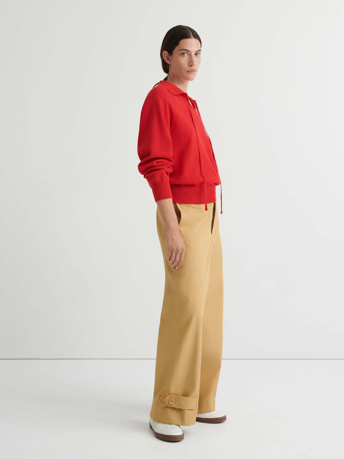 Person wearing a relaxed fit Keyhole Jumper – Poppy Red, beige wide-leg pants, and white shoes stands sideways against a plain white background, showcasing effortless knitwear style.