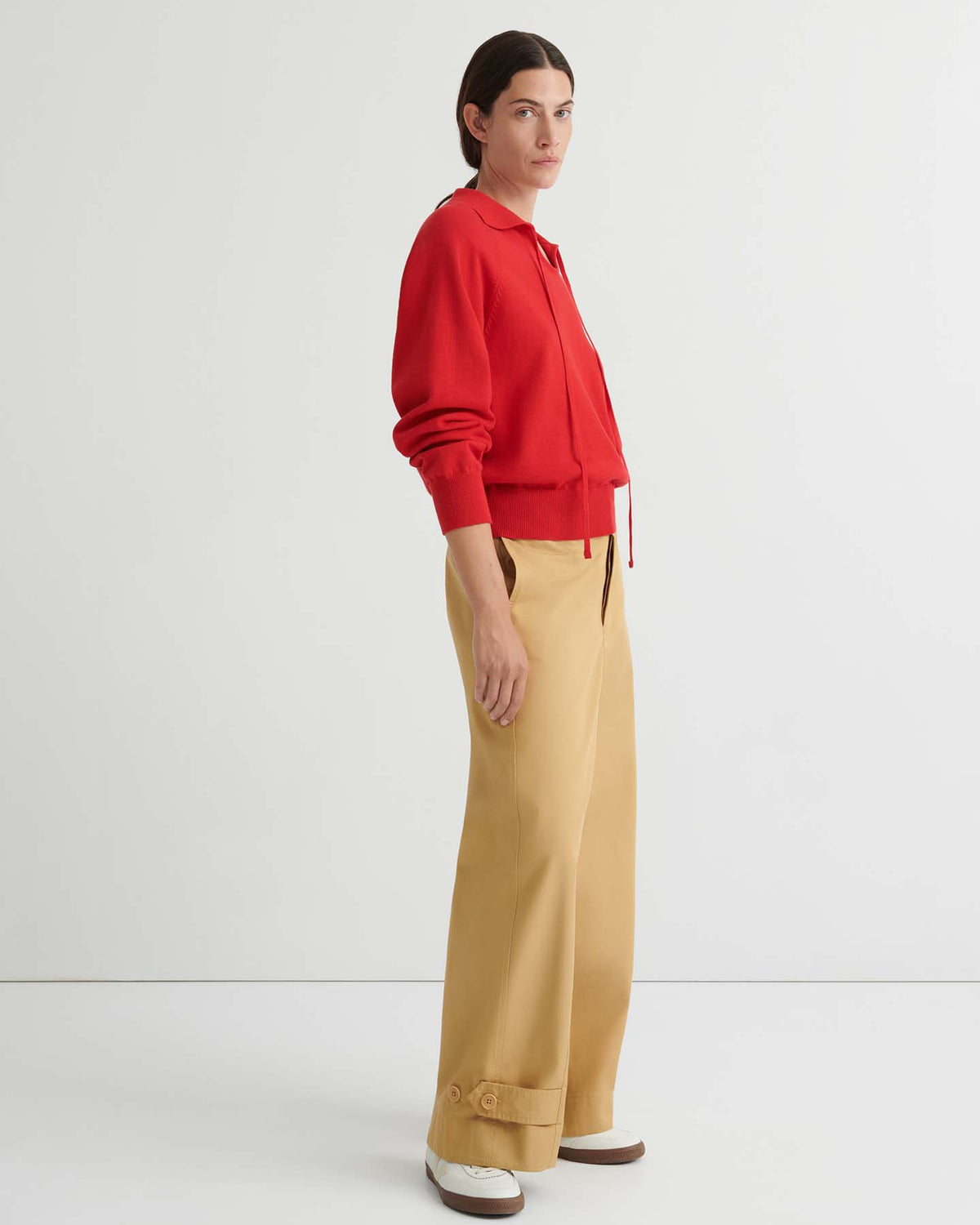 Person wearing a relaxed fit Keyhole Jumper – Poppy Red, beige wide-leg pants, and white shoes stands sideways against a plain white background, showcasing effortless knitwear style.