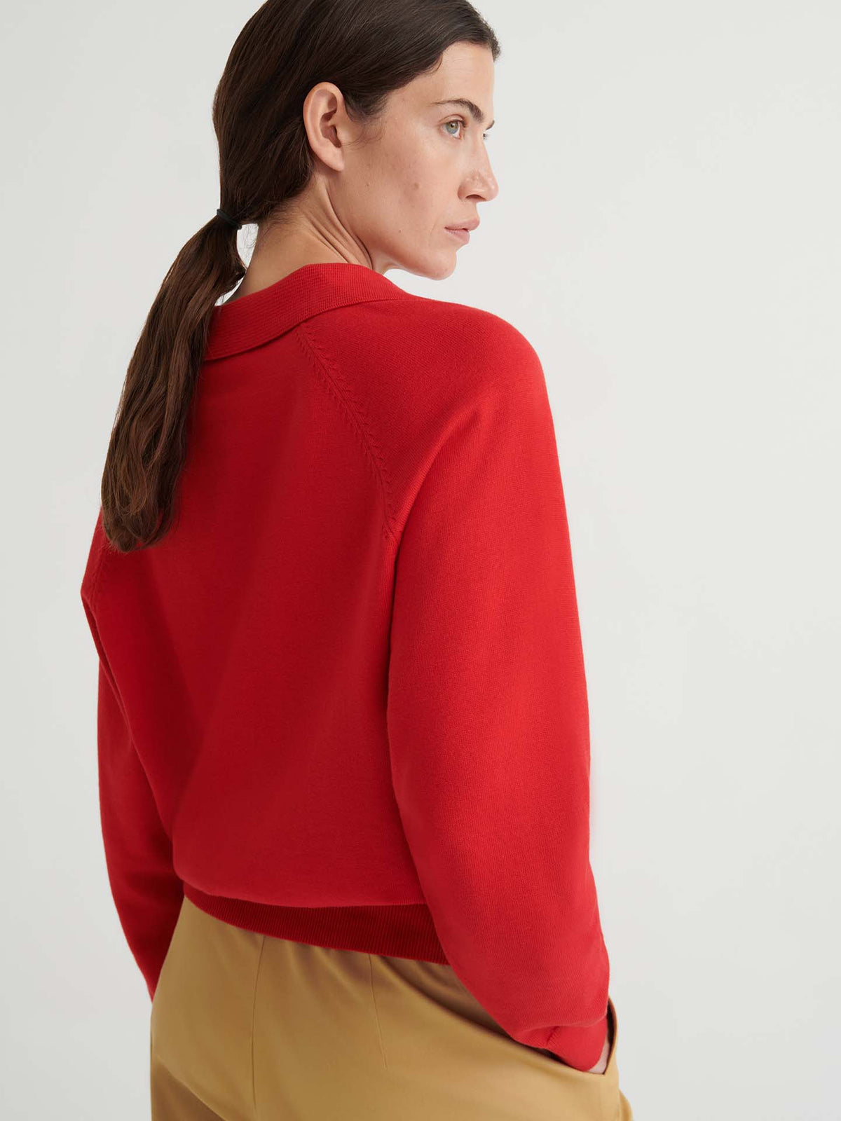 A woman with brown hair in a low ponytail wears the Keyhole Jumper in Poppy Red—a relaxed-fit knitwear top—paired with tan pants, standing against a plain light background.