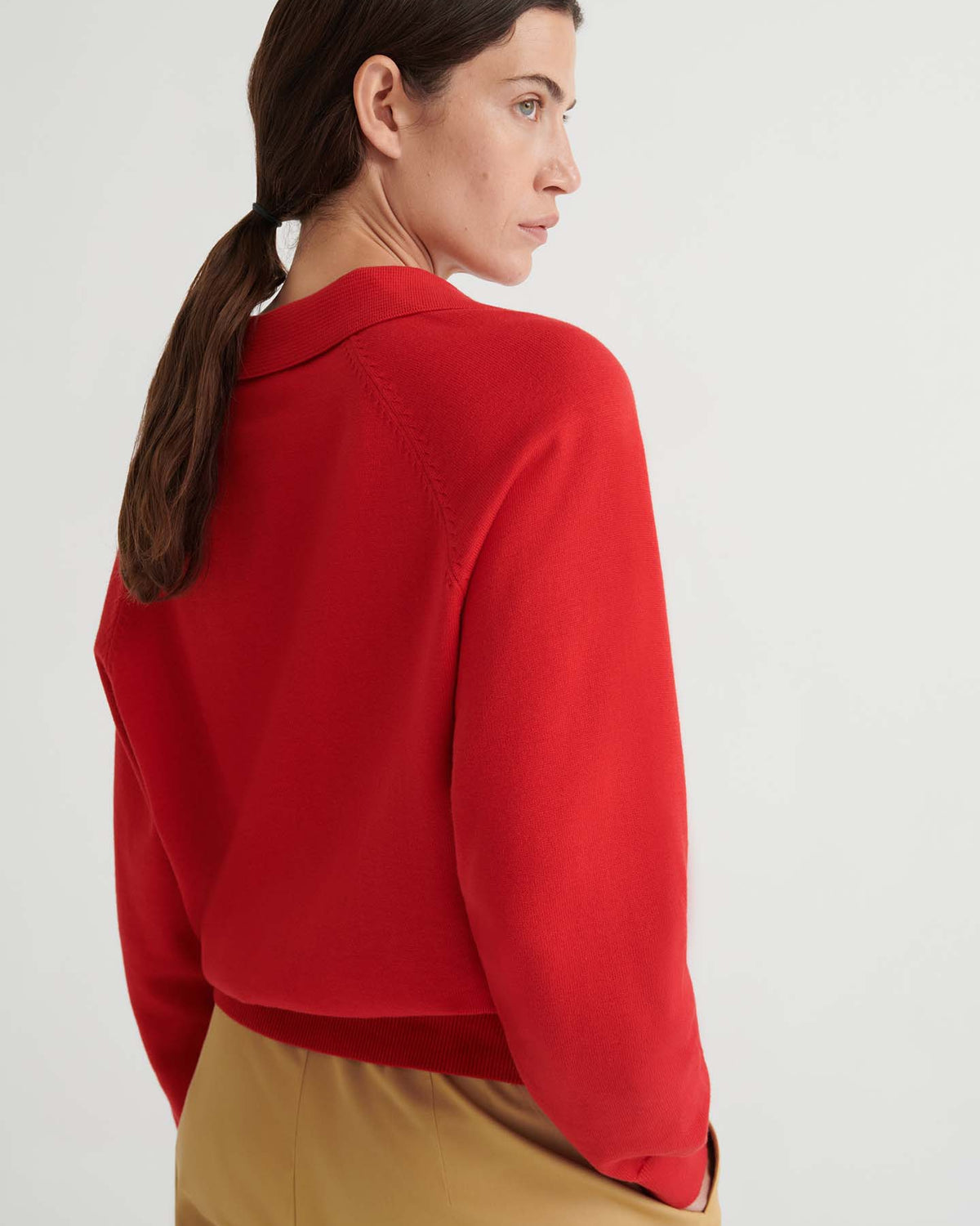 A woman with brown hair in a low ponytail wears the Keyhole Jumper in Poppy Red—a relaxed-fit knitwear top—paired with tan pants, standing against a plain light background.