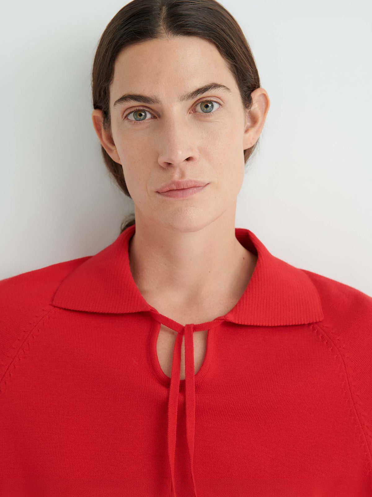 A woman with dark hair pulled back, wearing the Keyhole Jumper – Poppy Red featuring a front tie and relaxed fit, looks directly at the camera against a plain light background.