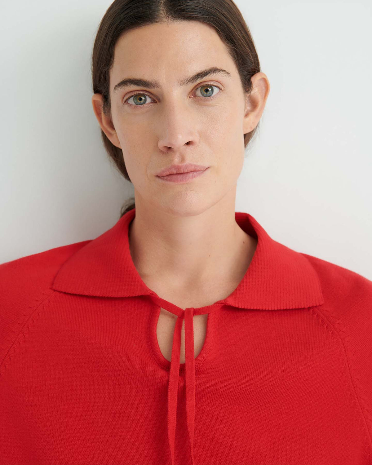 A woman with dark hair pulled back, wearing the Keyhole Jumper – Poppy Red featuring a front tie and relaxed fit, looks directly at the camera against a plain light background.