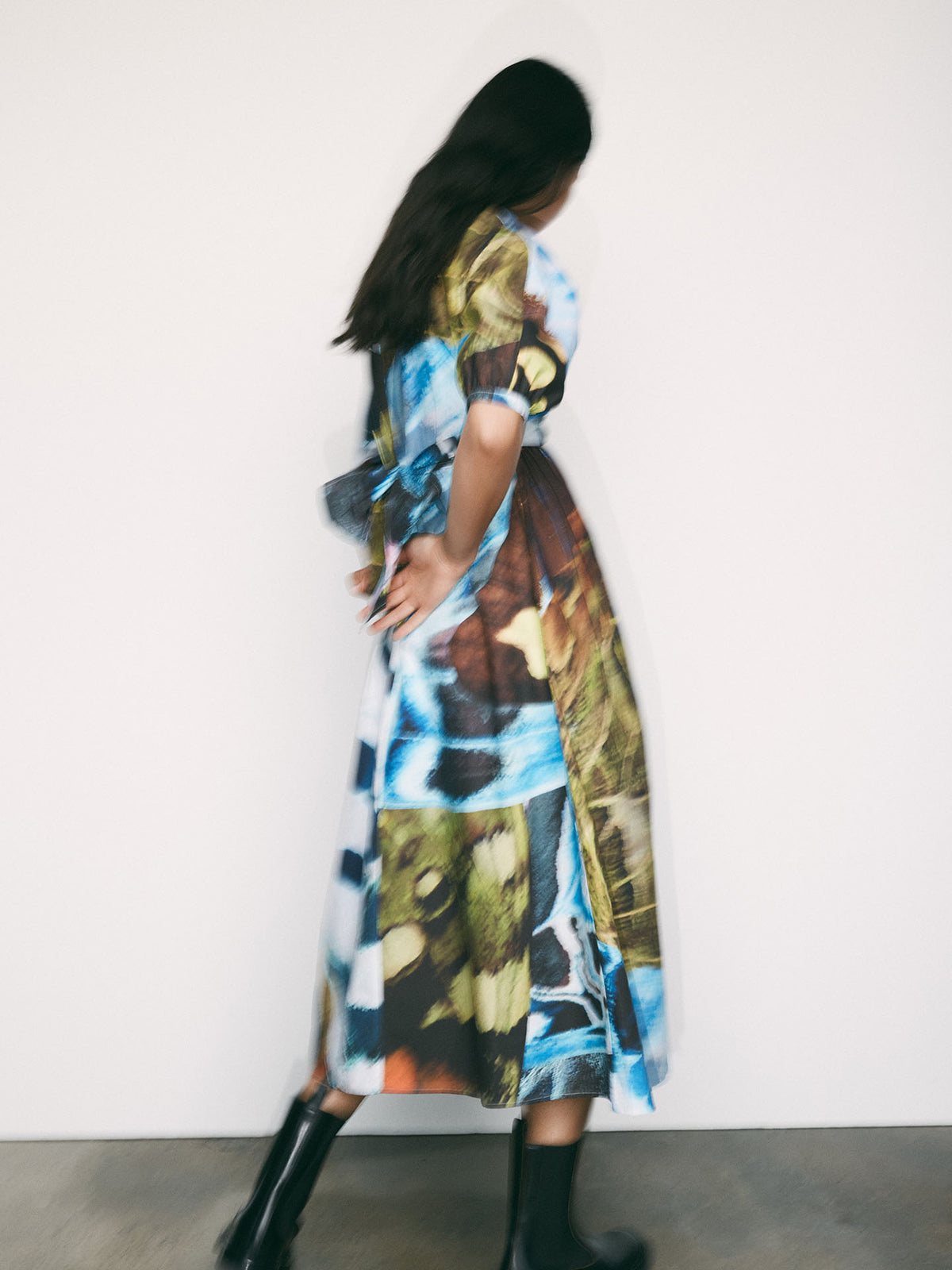 A woman wearing the Kowtow Marta Dress – Monarch, featuring a colorful abstract print and adjustable wrap tie, stands with her back to a plain white wall, hands clasped behind her, paired with black boots.