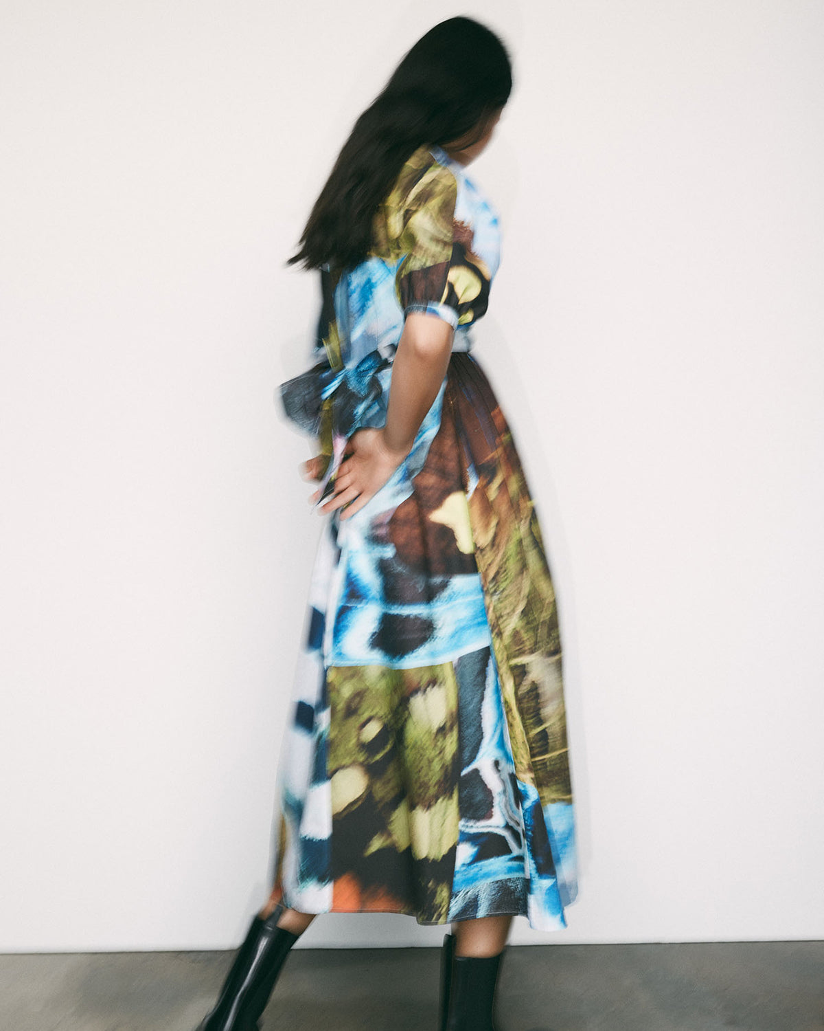 A woman wearing the Kowtow Marta Dress – Monarch, featuring a colorful abstract print and adjustable wrap tie, stands with her back to a plain white wall, hands clasped behind her, paired with black boots.