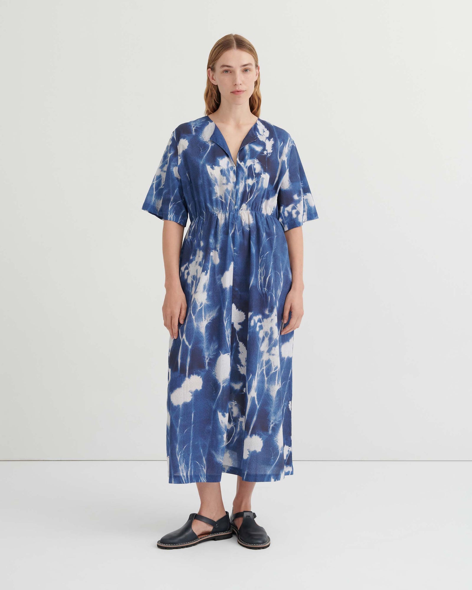 A woman stands against a plain white background wearing a blue and white tie-dye midi dress with short sleeves and black sandals.