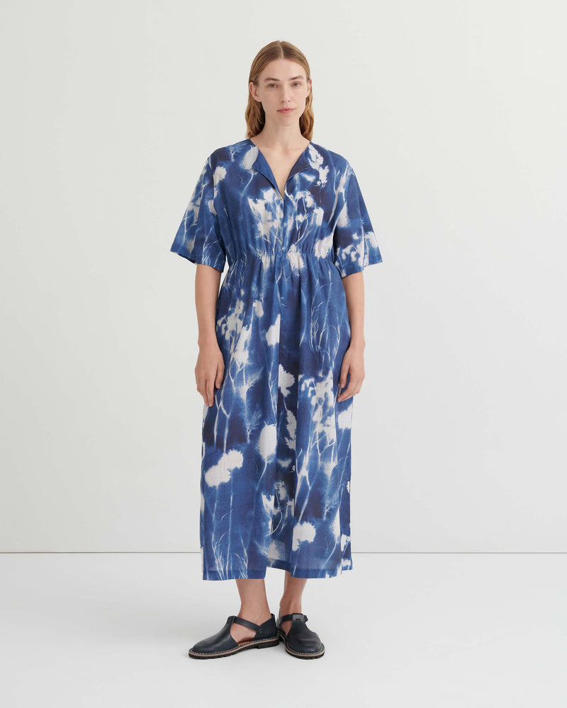 A woman stands against a plain white background wearing a blue and white tie-dye midi dress with short sleeves and black sandals.