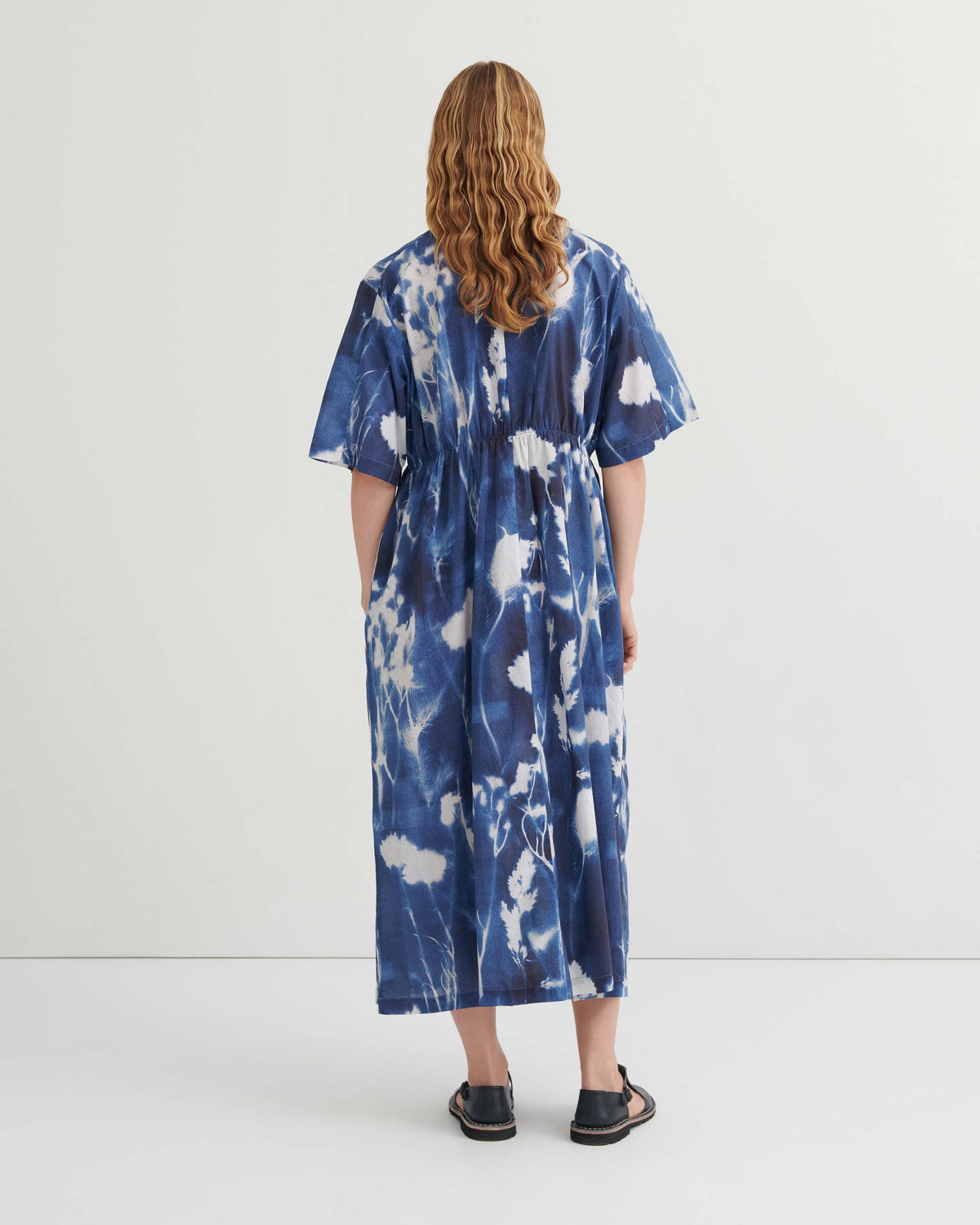 Person with wavy hair wears a blue and white tie-dye ankle-length dress and black sandals, standing with their back to the camera against a plain white background.