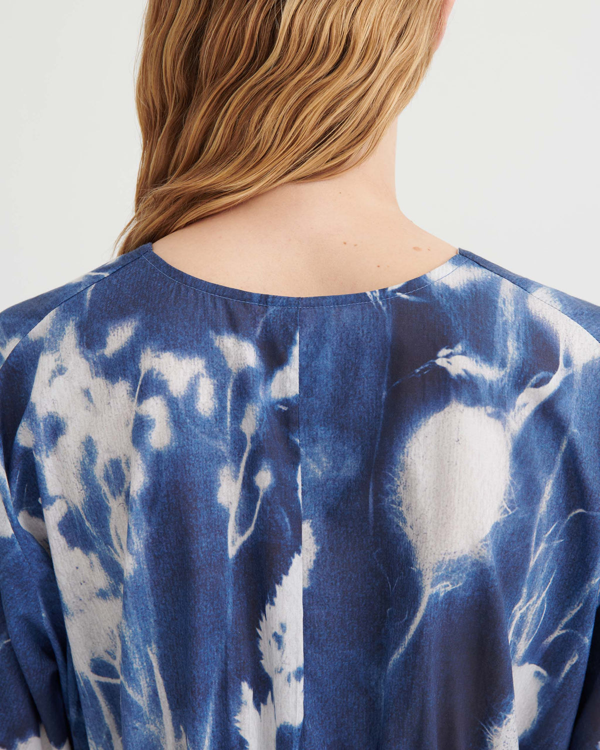 A person with wavy blonde hair is shown from behind, wearing a blue and white patterned top featuring abstract, tie-dye-like designs.