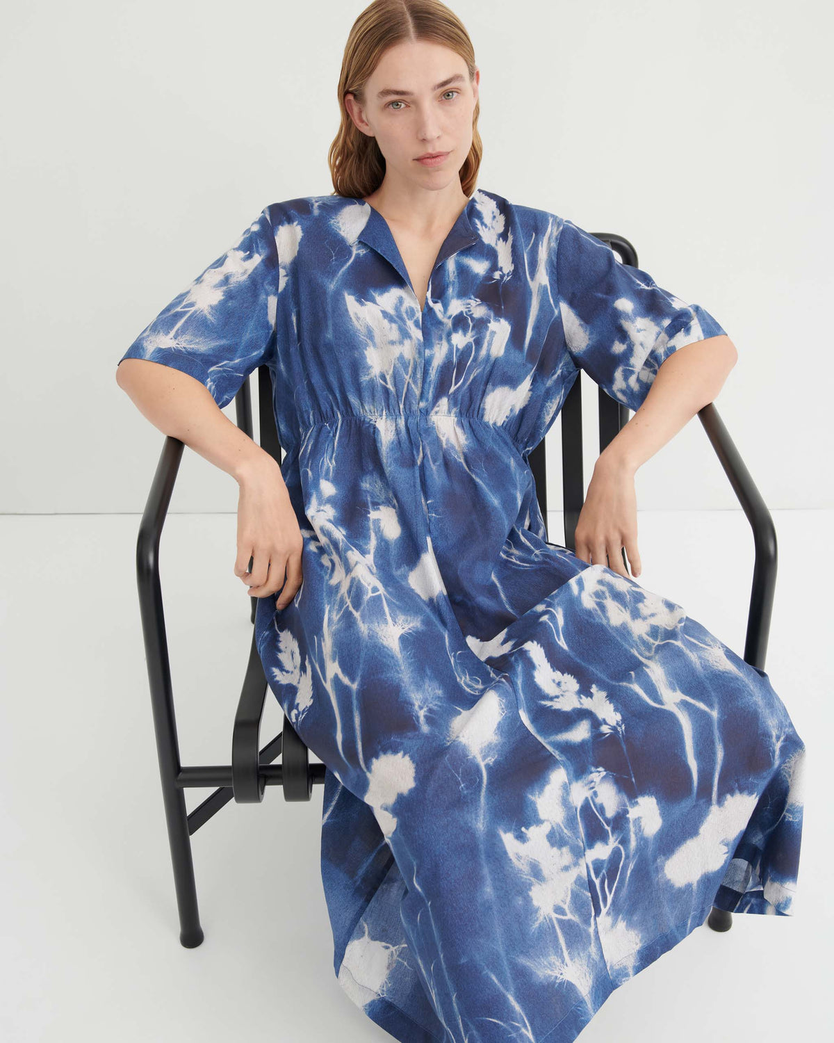 Woman sitting on a black chair, wearing a blue and white patterned dress, with a neutral expression, in a minimalist white room.