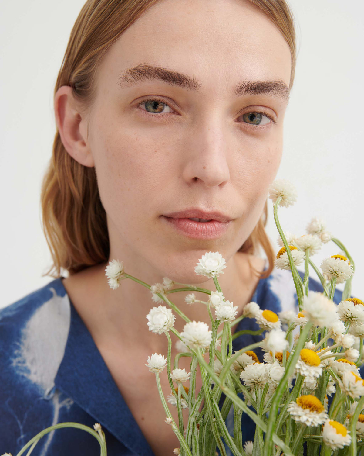 A person with light skin and straight, light brown hair holds a bouquet of white and yellow flowers, looking into the camera against a plain background.