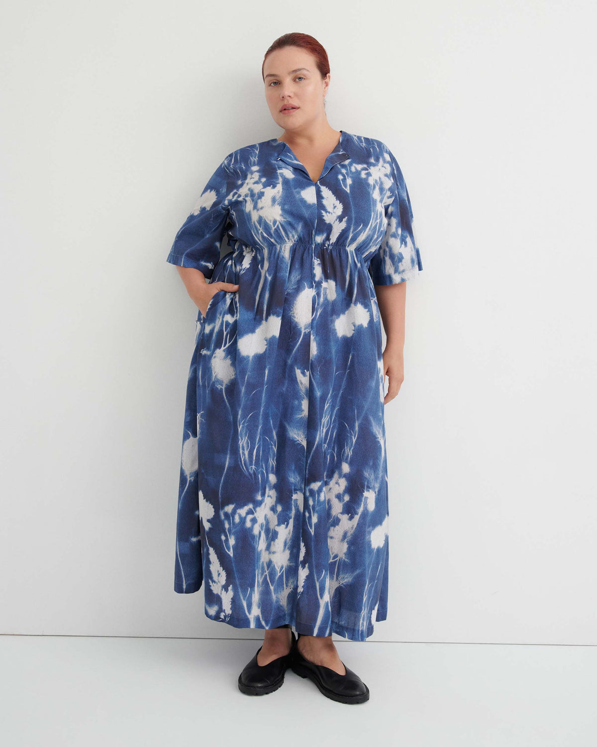 Person in a blue and white tie-dye maxi dress with short sleeves stands against a white wall, hands in pockets, wearing black flat shoes.