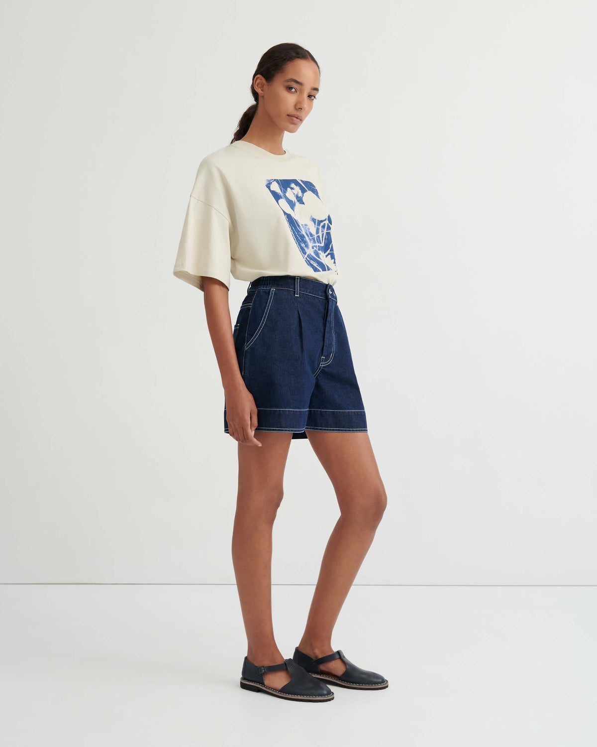 A person stands sideways wearing a white graphic T-shirt, dark denim shorts, and black flat shoes against a plain white background.