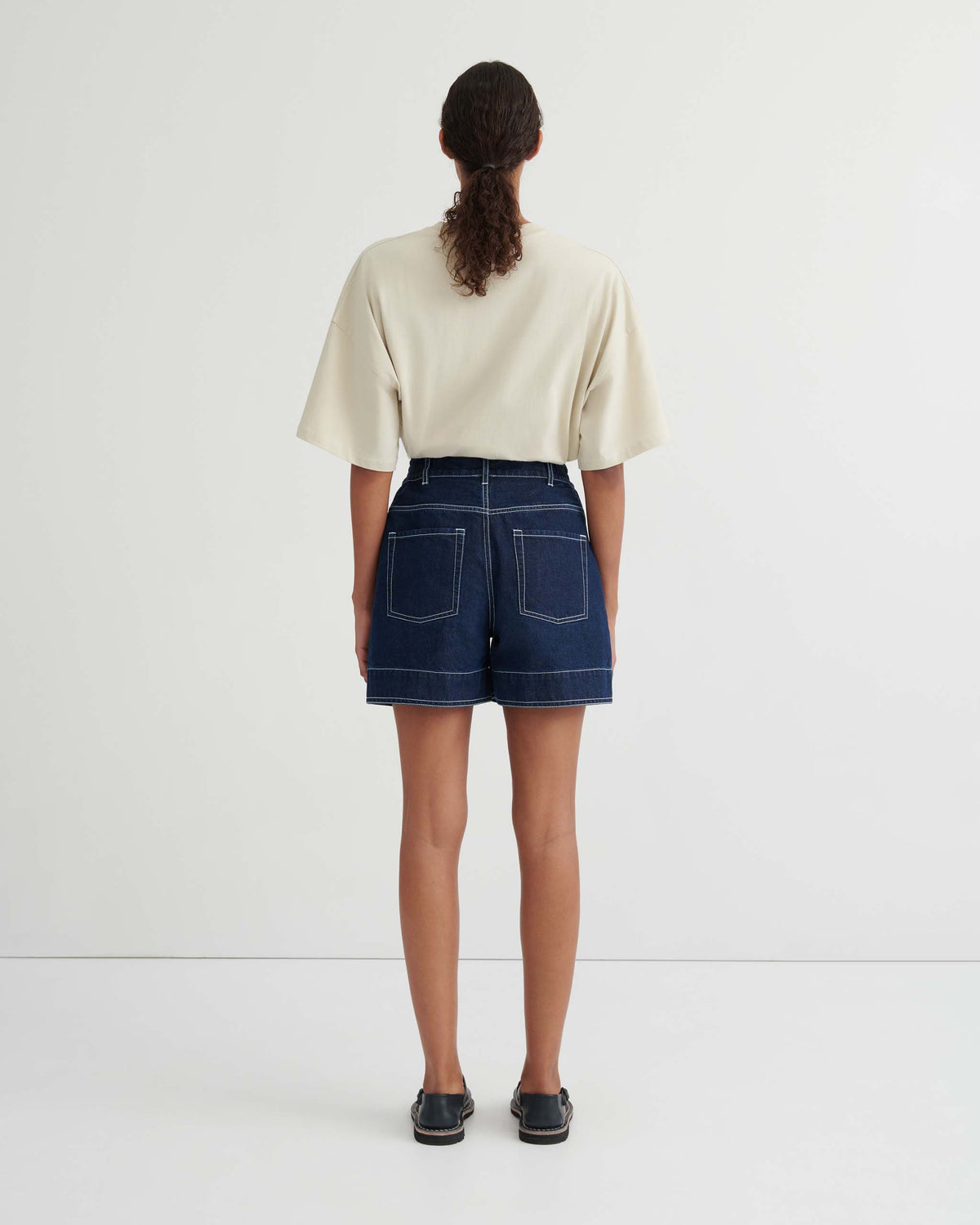 A person stands facing away, wearing a beige short-sleeve top, dark denim shorts with white stitching, and black shoes, against a plain white background.