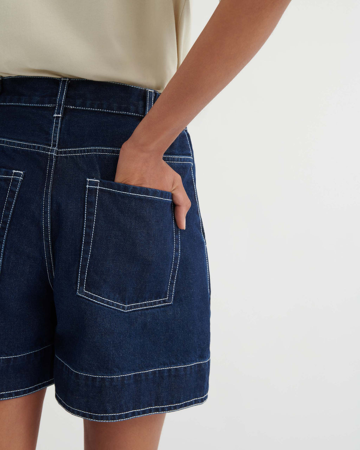 Person wearing dark denim shorts with white stitching, placing a hand in the back pocket. The background is plain and neutral.