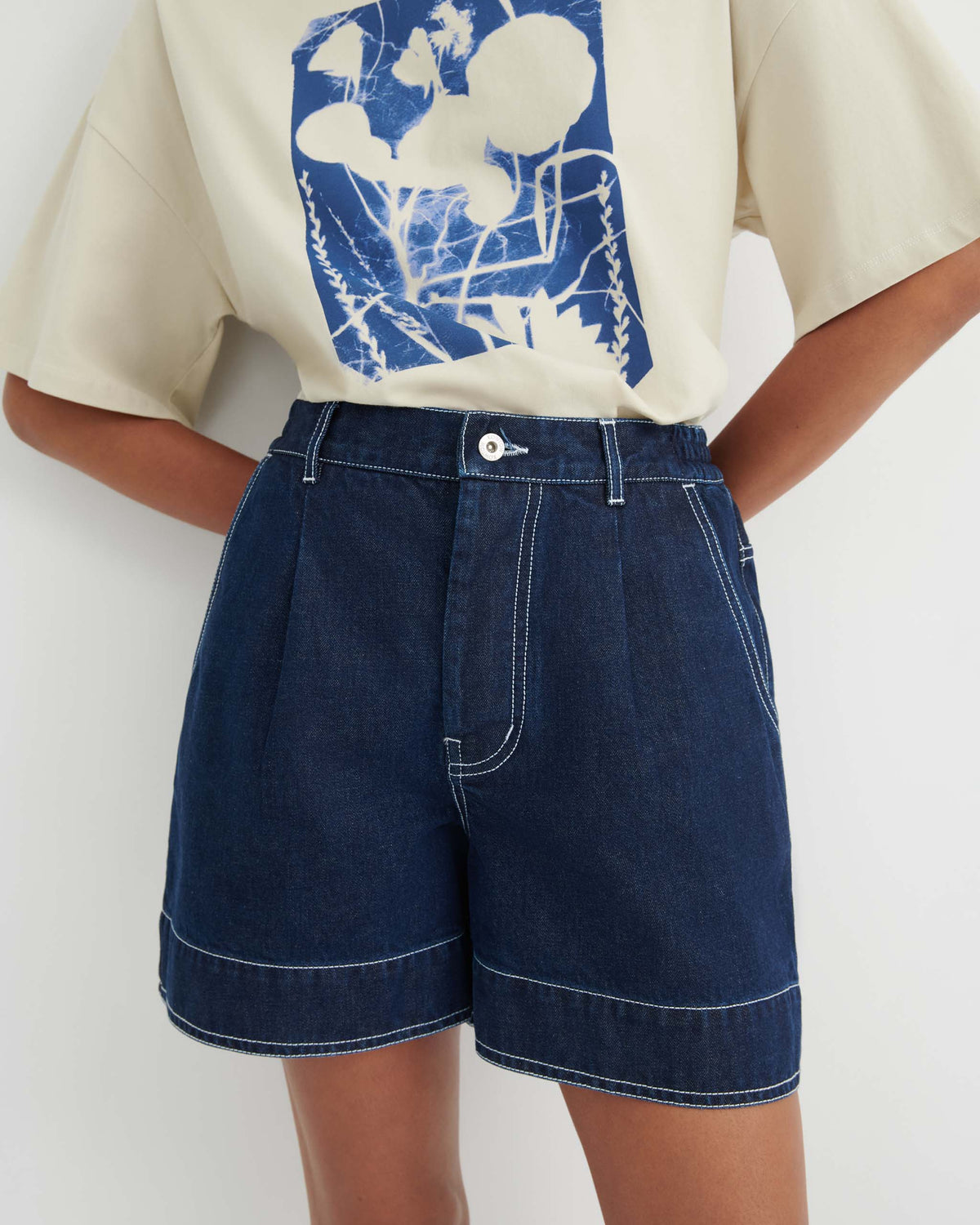 Person wearing high-waisted dark denim shorts with white stitching and a light t-shirt featuring a blue and white botanical graphic.