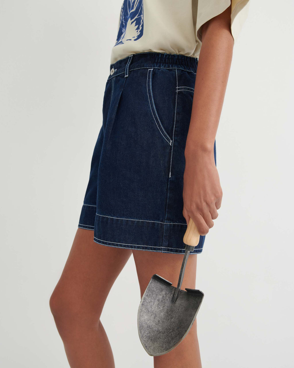 Person wearing a beige shirt and denim shorts holds a small gardening shovel with a wooden handle in their right hand against a plain background.