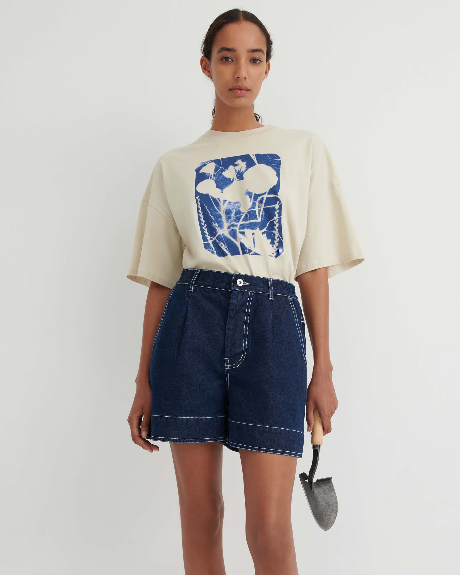 Woman in a cream t-shirt with a blue floral graphic, dark denim shorts, and holding a small garden trowel, standing against a plain white background.