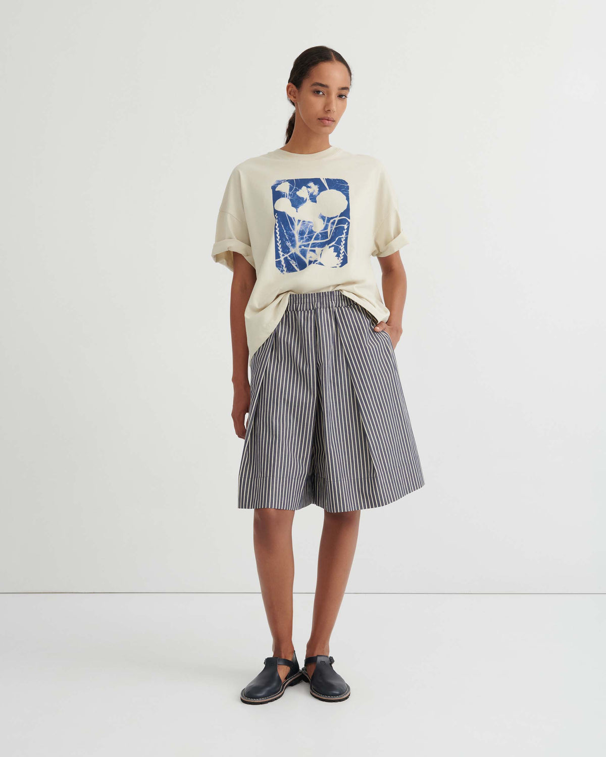 A person stands against a white wall wearing a cream graphic T-shirt, loose striped knee-length shorts, and black sandals.