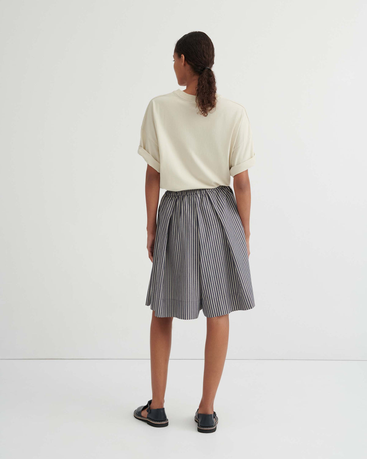 Person standing facing away, wearing a short-sleeve cream shirt, a black-and-white striped skirt, and black sandals against a plain white background.