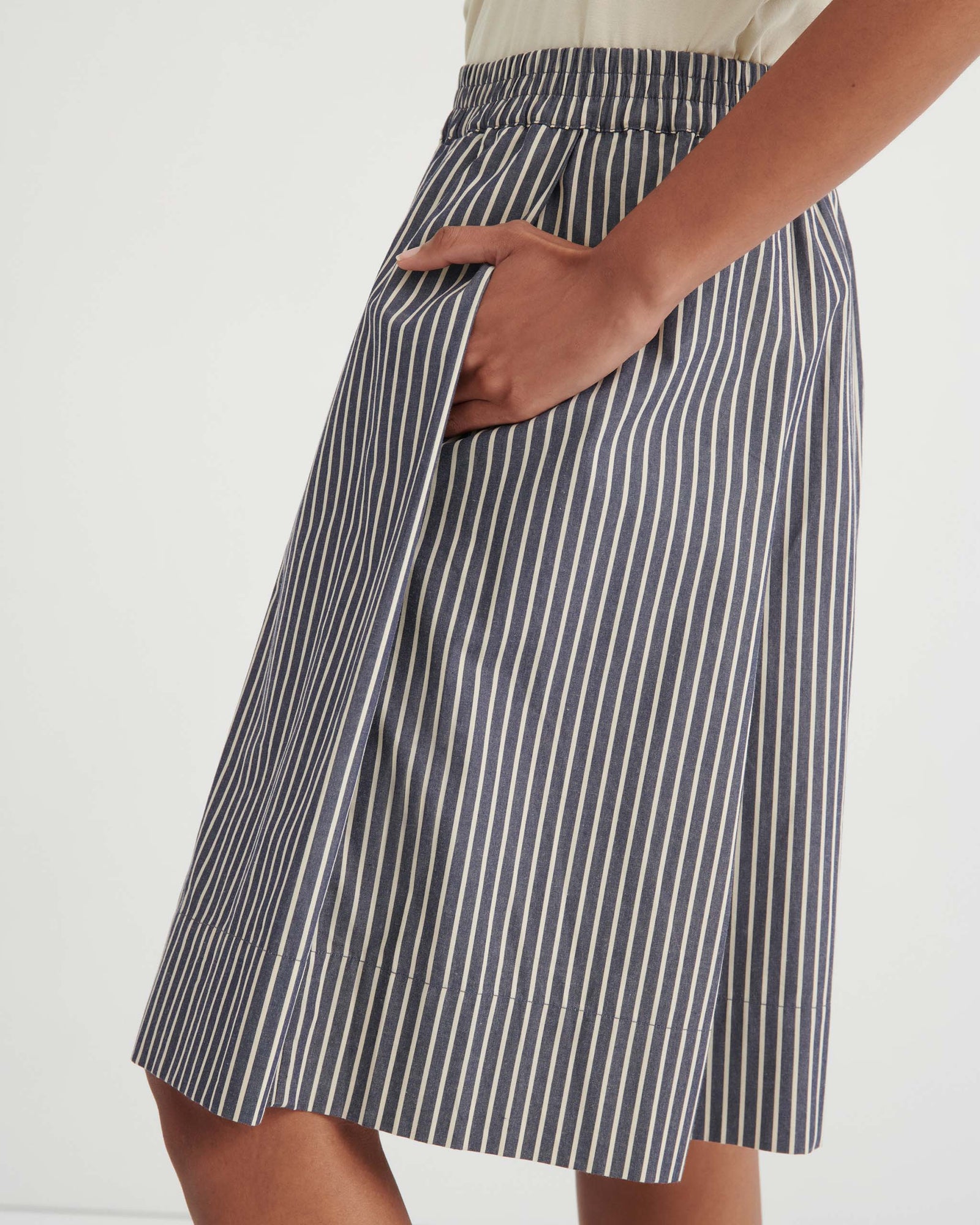 A person stands against a white wall wearing a cream graphic t-shirt, blue and white striped knee-length shorts, and black slip-on shoes.