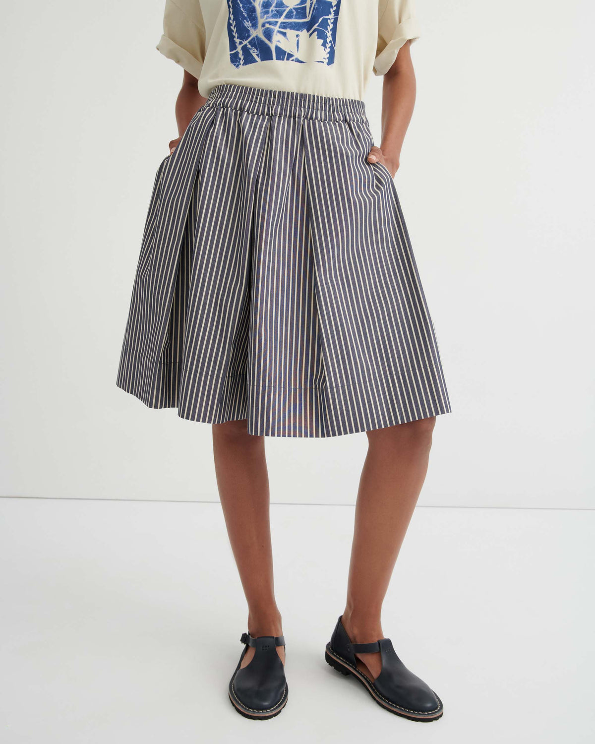 Person wearing a cream graphic T-shirt, a knee-length navy and white striped skirt, and black shoes stands against a plain white background.