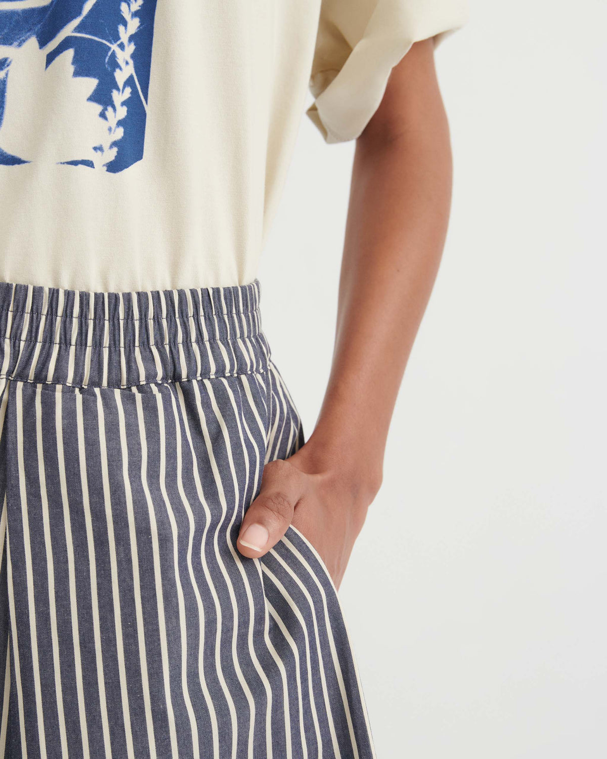 A person stands with their hand in the pocket of blue and white vertically striped pants, wearing a cream shirt with a blue graphic.