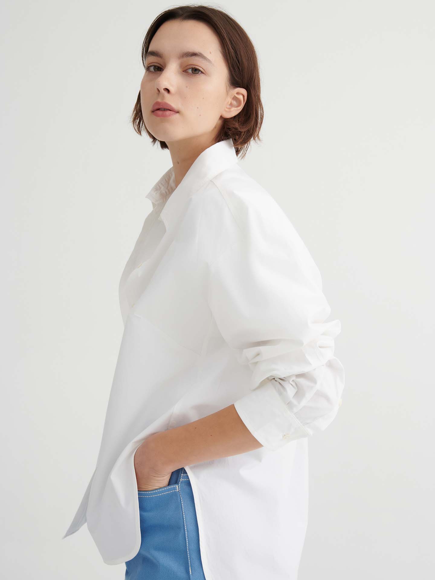 A person with short brown hair wears a Kowtow Season Shirt in white, styled in a relaxed fit with blue pants, standing against a plain white background and facing slightly sideways.